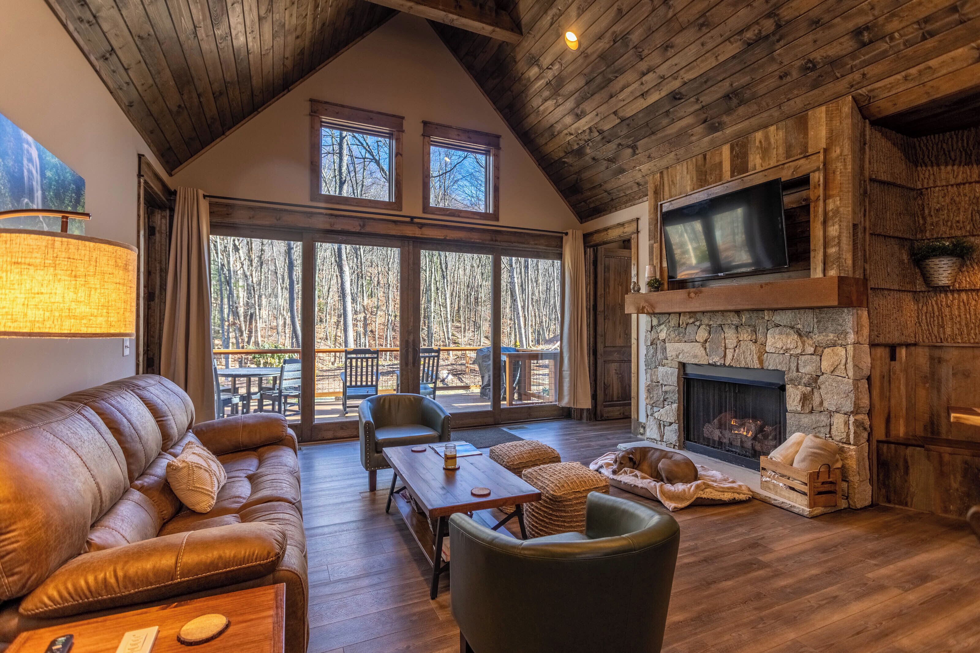 Interior acogedor de una cabaña con chimenea de piedra, sofá de cuero y un amplio ventanal con vistas a un bosque.