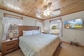 Une chambre avec un plafond en bois, un lit recouvert d’une courtepointe blanche, deux fenêtres avec des rideaux, une table de chevet, une lampe de chevet et un téléviseur fixé au mur.