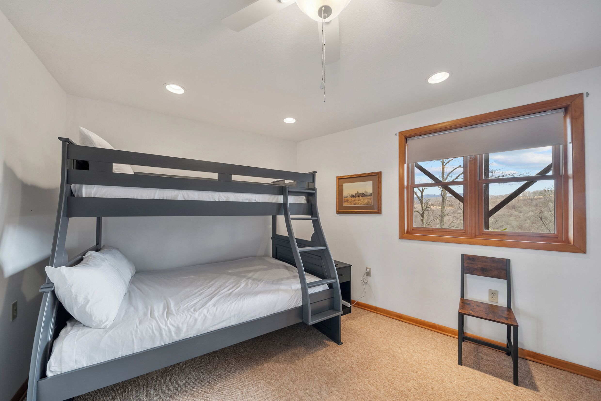 A bunk bed with a ladder, a small desk, a chair, a window with a view, and a ceiling fan.