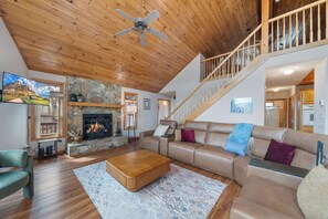 A spacious living room with a stone fireplace, wooden ceiling, and a staircase leading to an upper level.