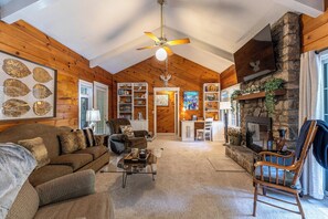 A cozy living room with a fireplace, wooden walls, a ceiling fan, and a bookshelf. - Creek House (Boone)