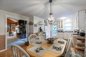 A dining area with a wooden table, white chairs, a chandelier, and a kitchen in the background. - Creek House (Boone)