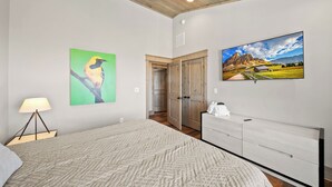 A bedroom with a bed, a nightstand, a lamp, a dresser, a painting of a bird, a painting of a mountain landscape, and a door leading to another room. - Zion at Eagles Nest (Banner Elk)