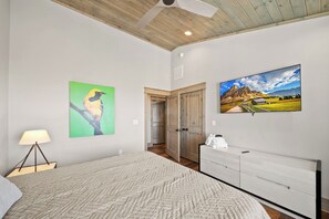 A bedroom with a bed, a nightstand, a lamp, a dresser, a painting of a bird, a painting of a mountain landscape, and a door leading to another room. - Zion at Eagles Nest (Banner Elk)