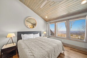 Une chambre à coucher avec un grand lit, un miroir rond, une table de chevet, une lampe et une fenêtre donnant sur un beau paysage.