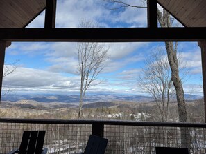 Une vue depuis un espace abrité, doté d’un plafond en bois et d’une rambarde en métal, donnant sur un paysage composé d’arbres et de montagnes lointaines.