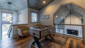 A spacious room with a foosball table, wooden flooring, a fireplace, and a view of the outdoors. - Zion at Eagles Nest (Banner Elk)