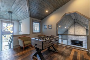 A spacious room with a foosball table, wooden flooring, a fireplace, and a view of the outdoors. - Zion at Eagles Nest (Banner Elk)
