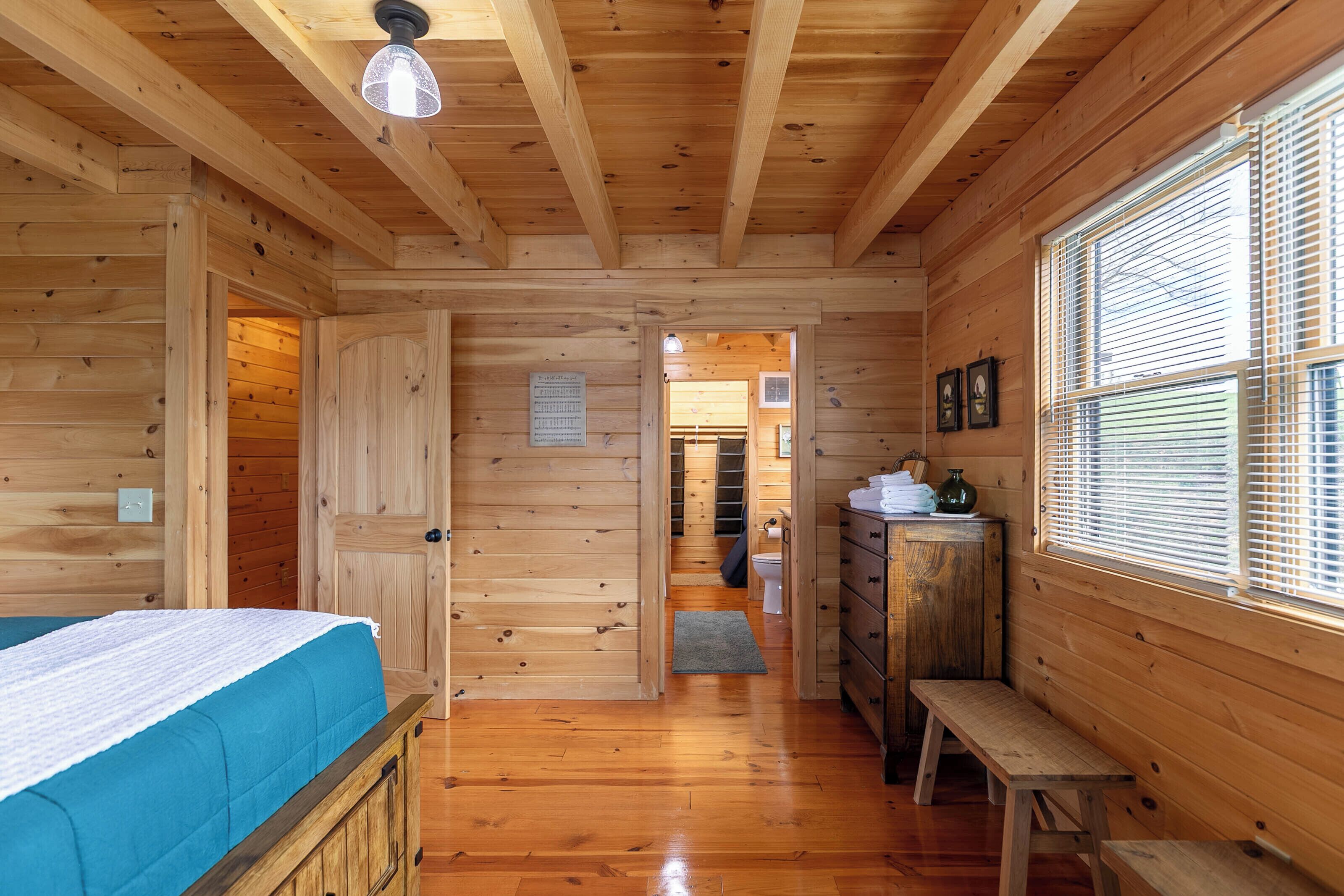 Interior de una cabaña de madera con una cama, un banco, un aparador y una ventana con persianas.