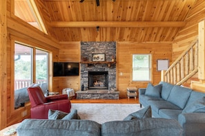 A log cabin interior with a stone fireplace, a red leather chair, a gray sofa, and a wooden staircase. - Chasing Clouds Cabin (Fleetwood)