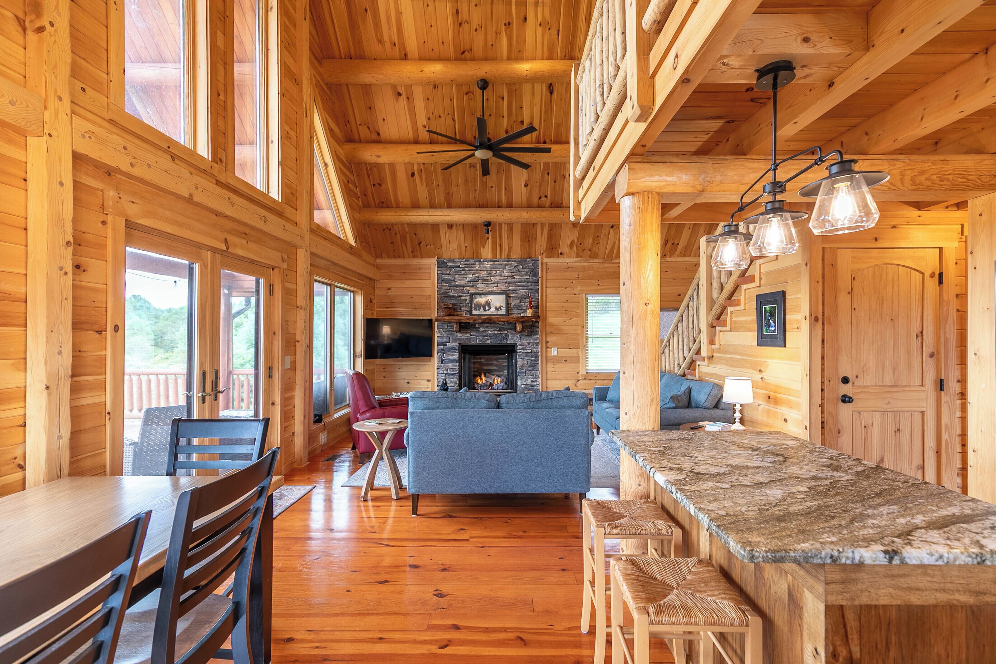 A log cabin interior with a stone fireplace, wooden dining table, and a bar area.
