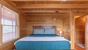 A wooden cabin bedroom with a bed, pillows, a nightstand, and a window with blinds. - Chasing Clouds Cabin (Fleetwood)