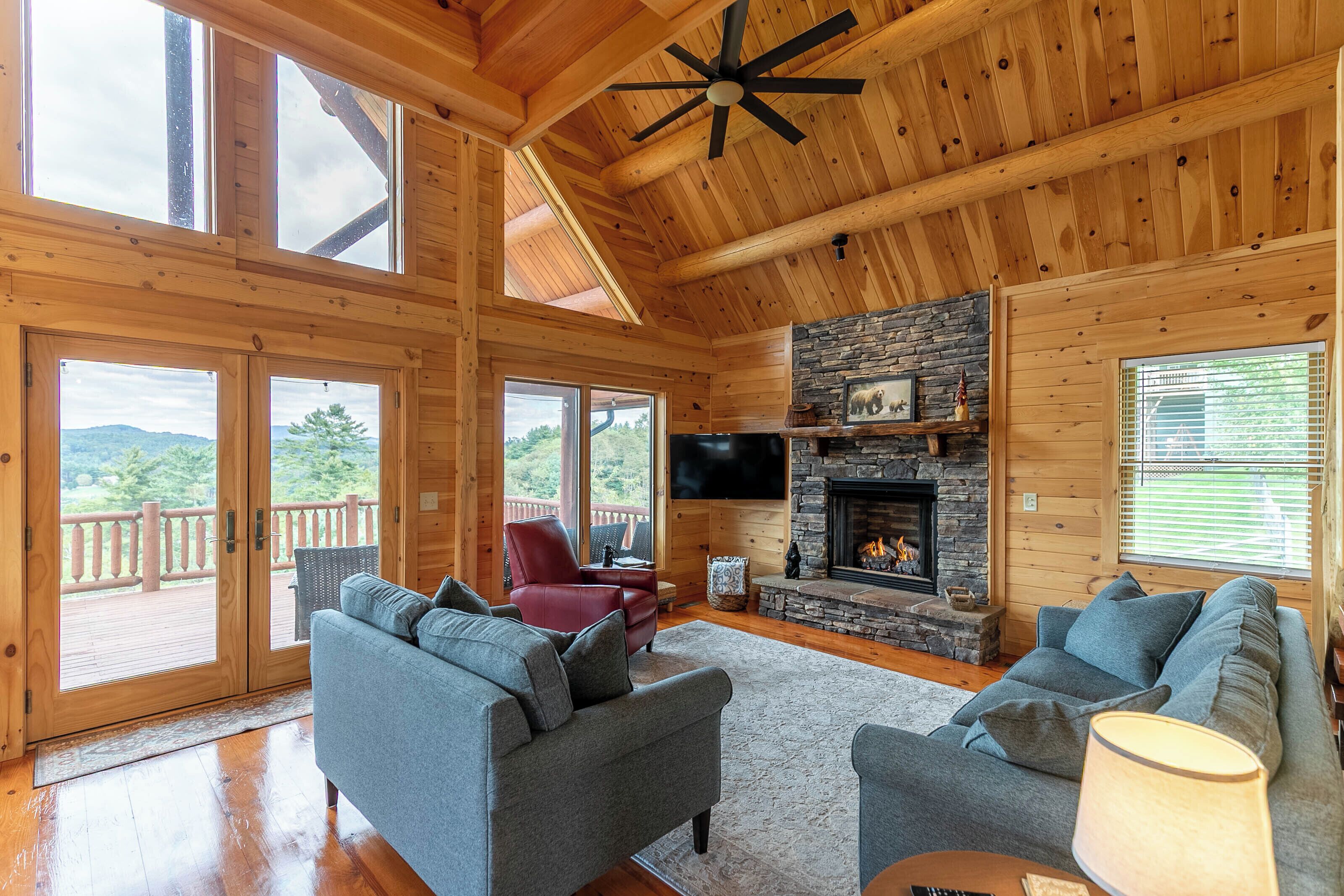 A spacious cabin interior with a stone fireplace, large windows, and a deck.