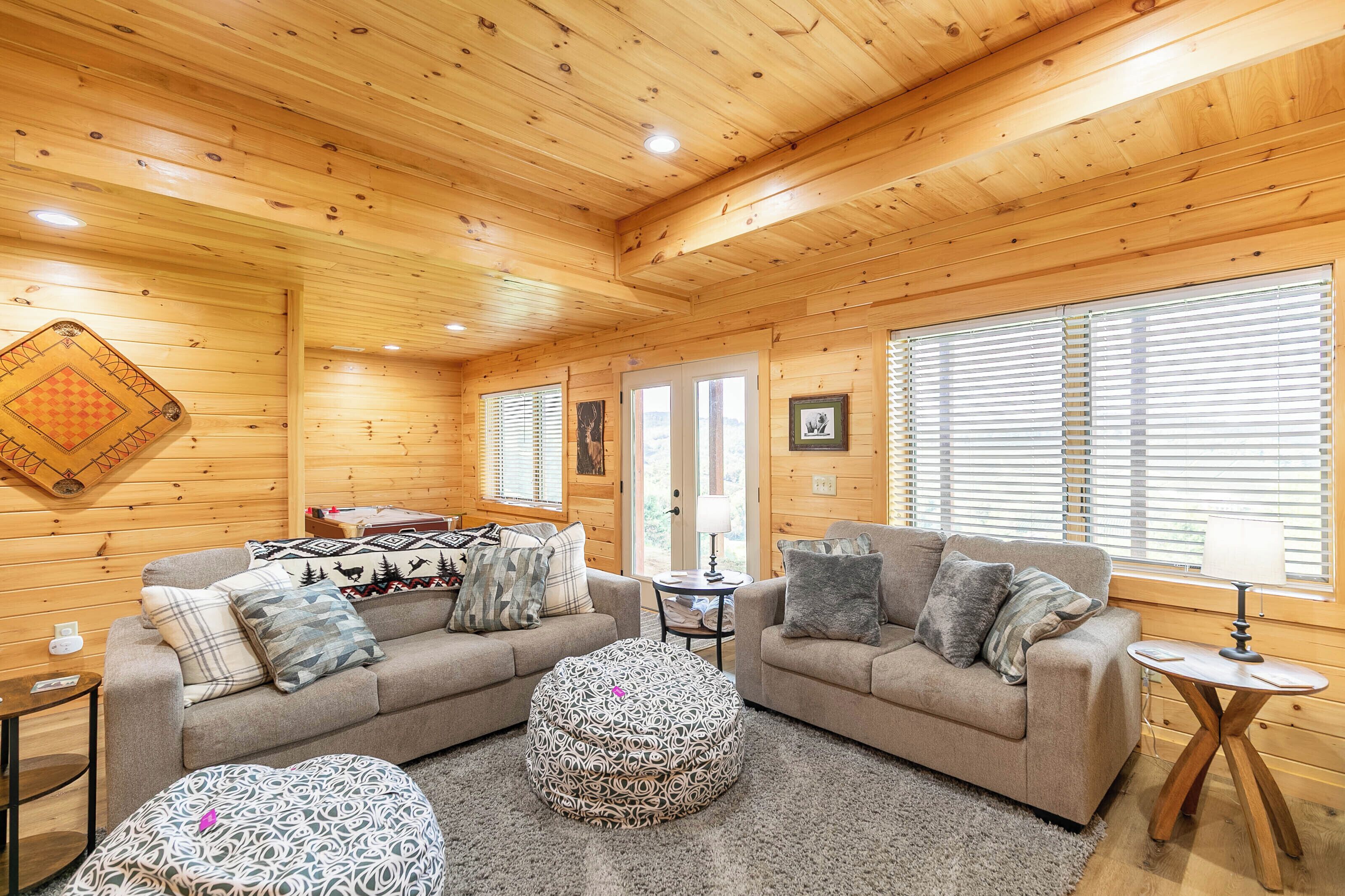 L’intérieur d’un chalet accueillant, avec un plafond en bois, un ensemble de canapés, des poufs, une petite table et une fenêtre avec des stores.