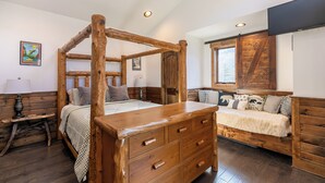 A four-poster bed with wooden headboard and drawers, a bedside table with a lamp, a bench with cushions, and a window with a view of trees. - Ripple Run at Eagles Nest (Banner Elk)