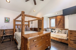 Ein Bett mit Baldachin, ein hölzernes Kopfende und Schubladen, ein Nachttisch mit Lampe, eine Bank mit Kissen und ein Fenster mit Blick auf Bäume.