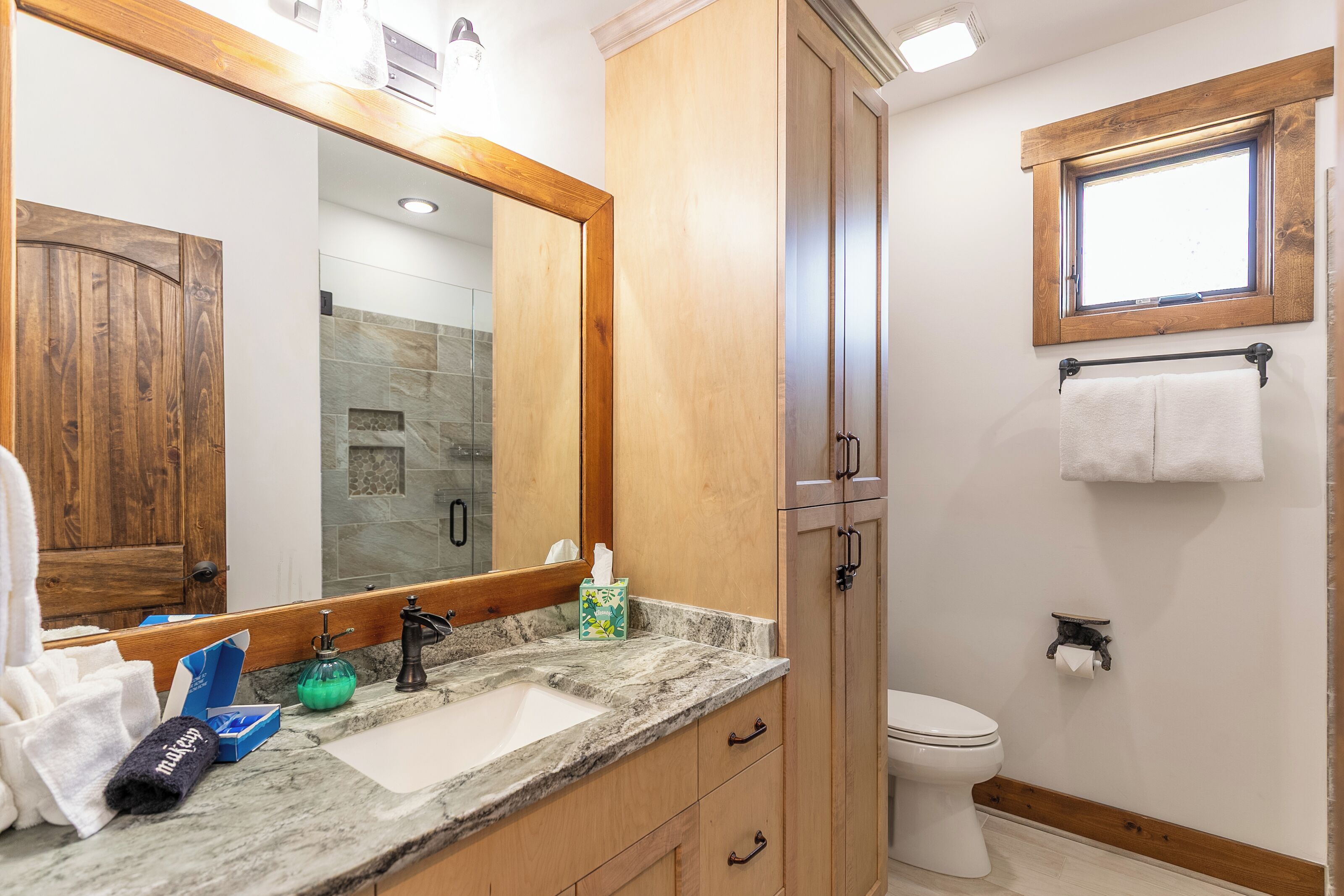 A bathroom with a wooden vanity, a large mirror, a marble countertop, a toilet, and a window.