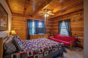 A cozy cabin room with a wooden interior, a bed with a quilt, a red sofa, a television on a wooden dresser, and a ceiling fan. - River Ridge (Boone)