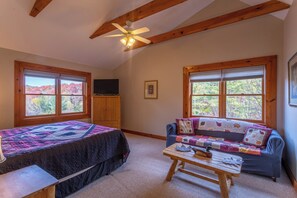 A cozy room with a bed, a sofa, a table, and a ceiling fan. There are large windows with a view of trees. - River Ridge (Boone)