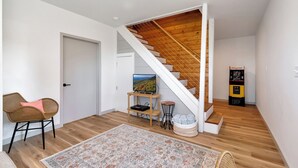 A modern interior with a wooden staircase, a flat-screen TV on a stand, a gaming console, and a wicker chair. - Calling Me Home (Lenoir)