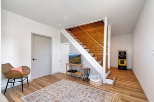 Un interior moderno con una escalera de madera, un televisor de pantalla plana sobre un mueble bajo, una consola de videojuegos y un sillón de mimbre.