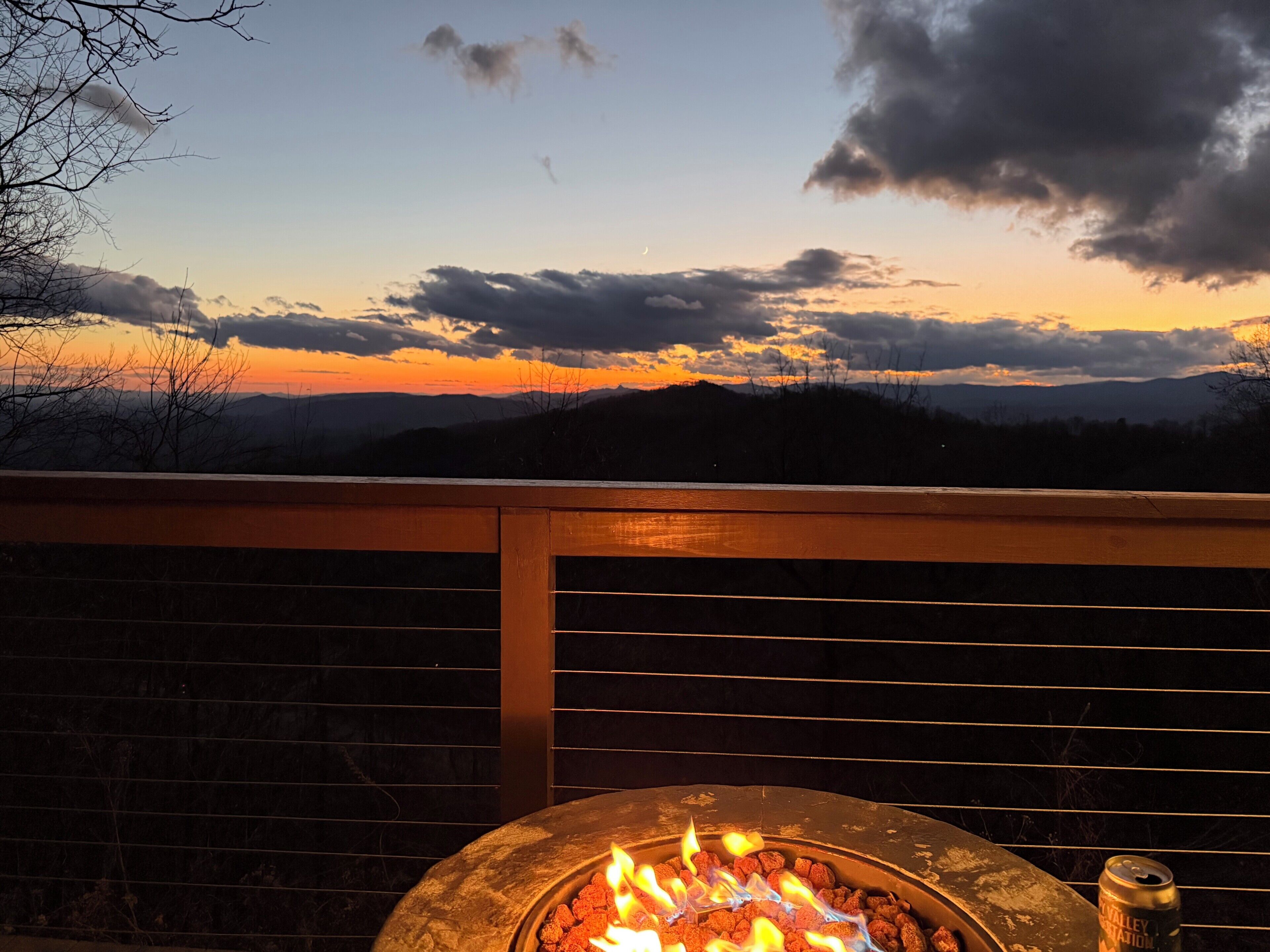 Ein Lagerfeuer mit Flammen, eine Bierdose und Blick auf einen Sonnenuntergang über Bergen.