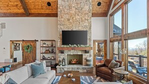 A spacious living room with a stone fireplace, a large TV mounted above it, and a view through large windows. - Altitude Adjustment at Eagles Nest (Elk Park)