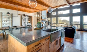 Una cocina amplia con una isla grande, armarios de madera y un salpicadero de piedra. Hay un comedor con mesa y sillas, y vistas al exterior a través de grandes ventanales.