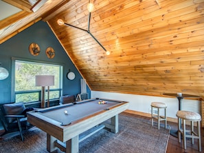 A cozy room with a pool table, wooden ceiling, and a window overlooking greenery.