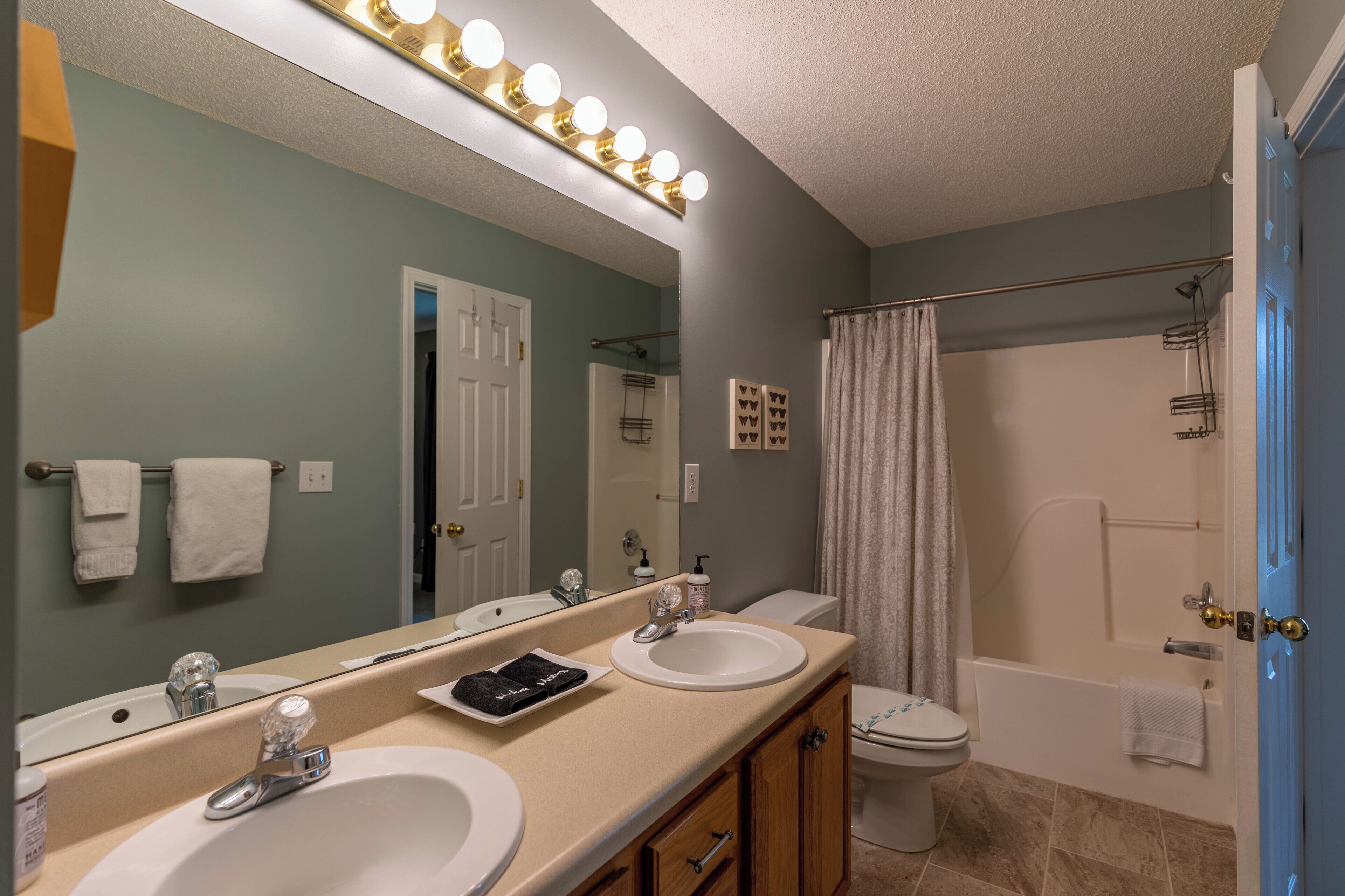 A bathroom with two sinks, a mirror with lights, a shower, and a toilet.