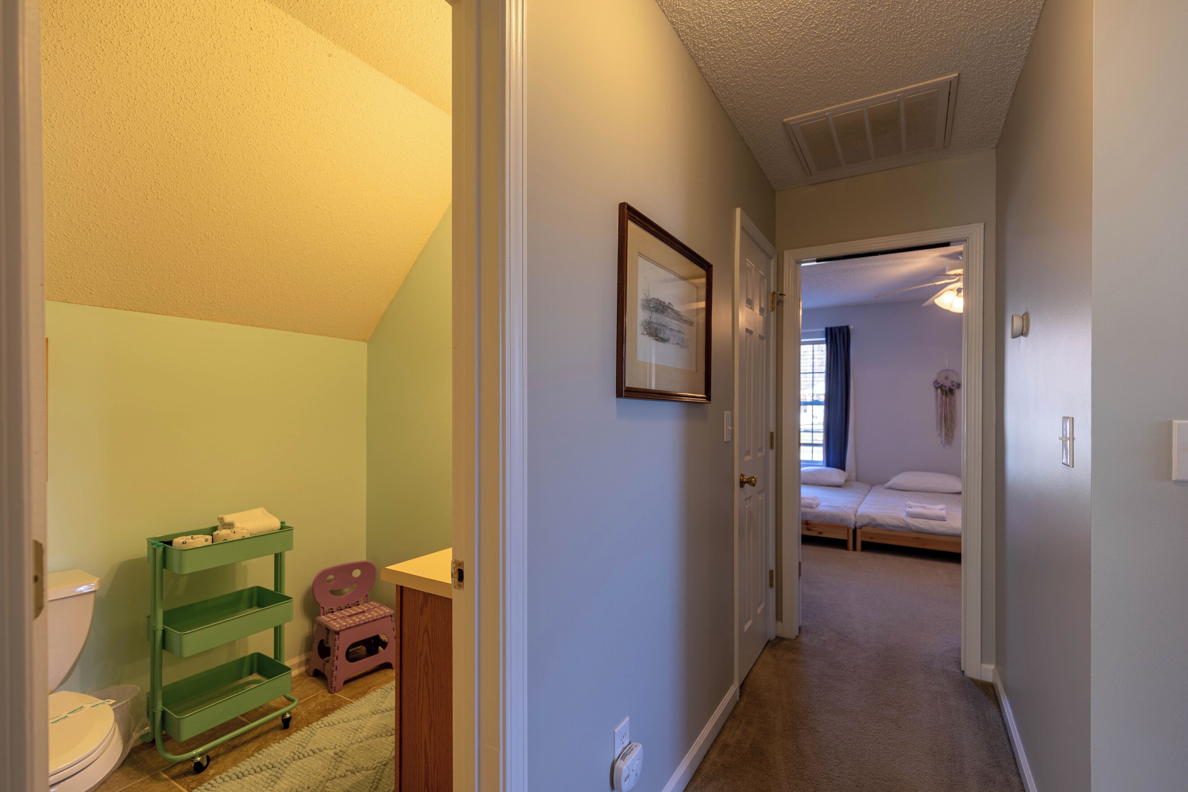 Un couloir avec un chariot de rangement vert, une petite chaise rose et un lit dans la pièce adjacente.