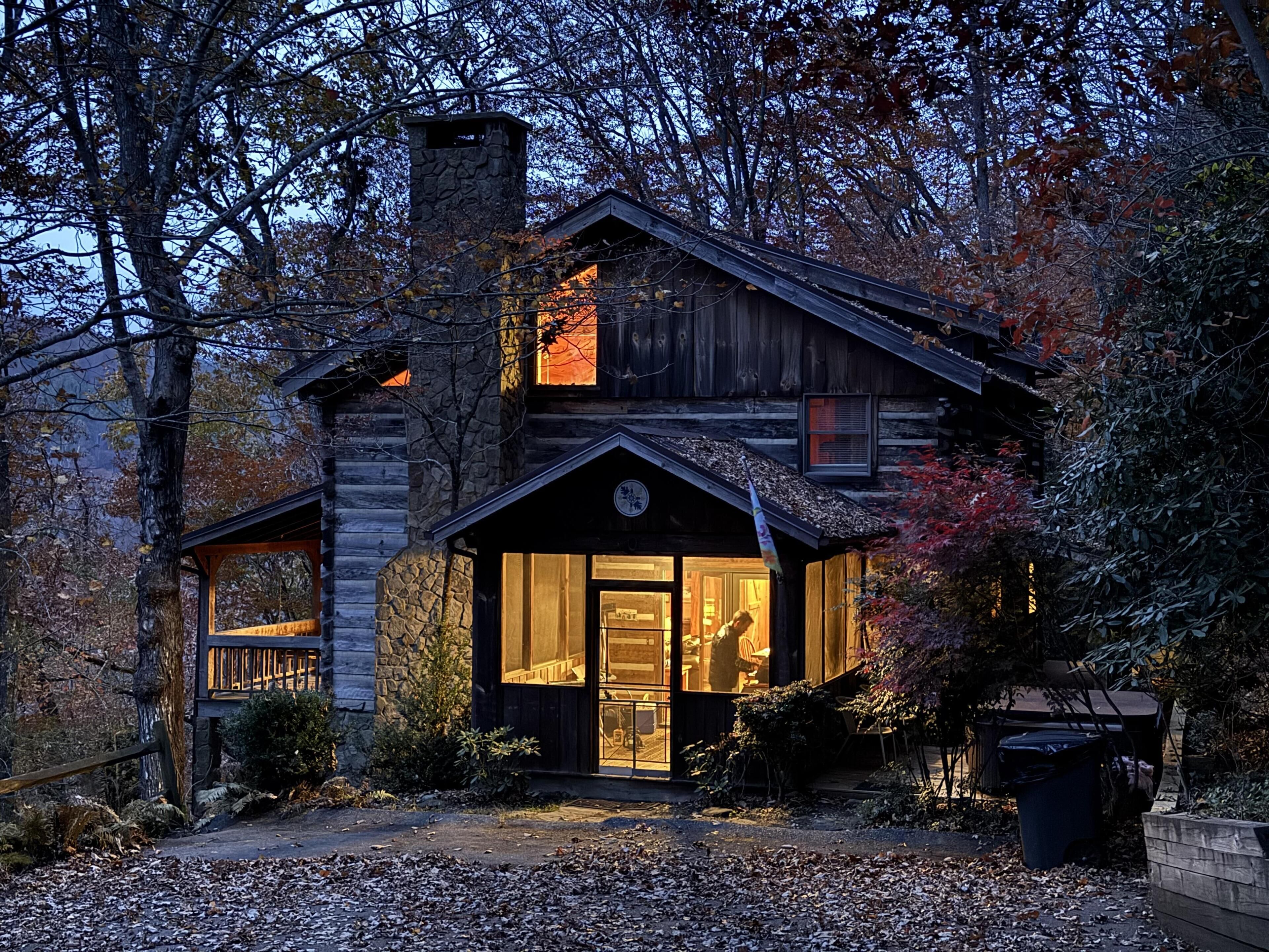 A rustic log cabin with a stone chimney, a large front window, and a wooden porch.