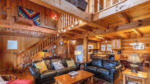 A log cabin interior with a living room featuring two black leather sofas, a wooden coffee table, and a red cushioned chair. A kitchen with wooden cabinets and a dining area with a wooden table and chairs is visible. - Daniel Boone Lodge (Boone)