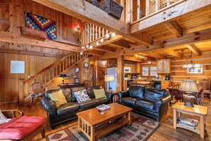 Ein Holzhaus-Interieur mit einem Wohnzimmer, ausgestattet mit zwei schwarzen Ledersofas, einem Holztisch und einem roten Sitzsessel. Eine Küche mit Holzschränken sowie ein Essbereich mit Holztisch und -stühlen sind ebenfalls zu sehen.