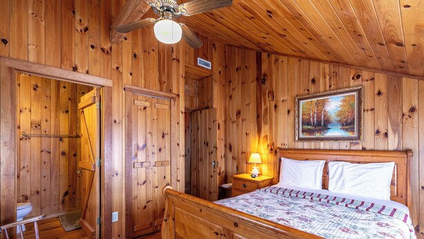 A wooden room with a bed, a nightstand with a lamp, a painting on the wall, and a ceiling fan. - Daniel Boone Lodge (Boone)