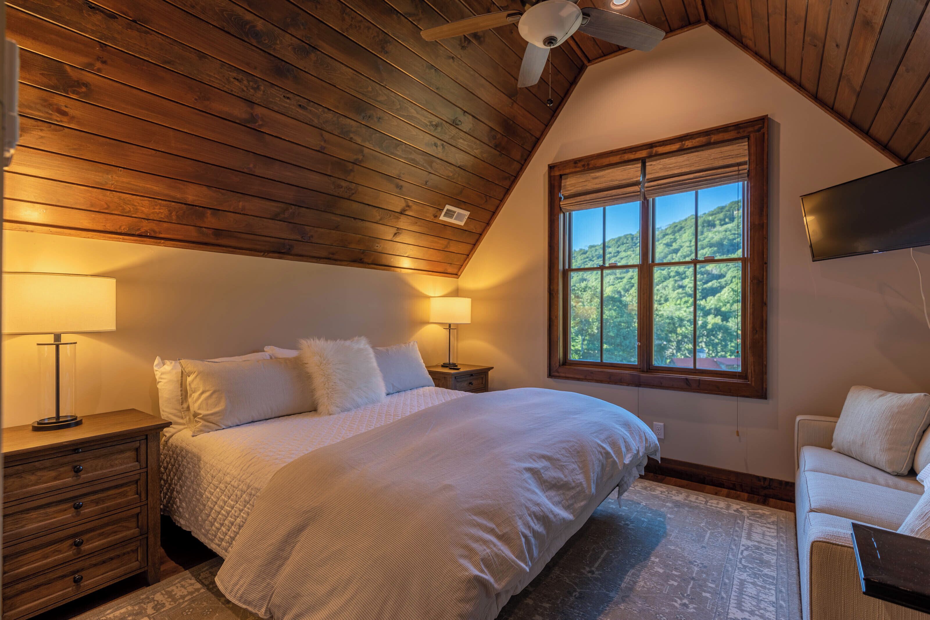 Un dormitorio con una cama grande, techo de madera, ventana con vista a follaje y televisor de pantalla plana montado en la pared.