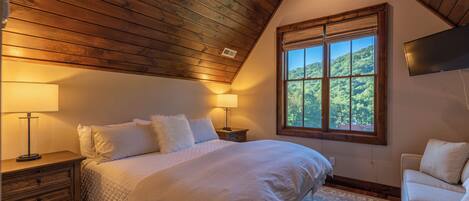 Un dormitorio con una cama grande, techo de madera, ventana con vista a follaje y televisor de pantalla plana montado en la pared.
