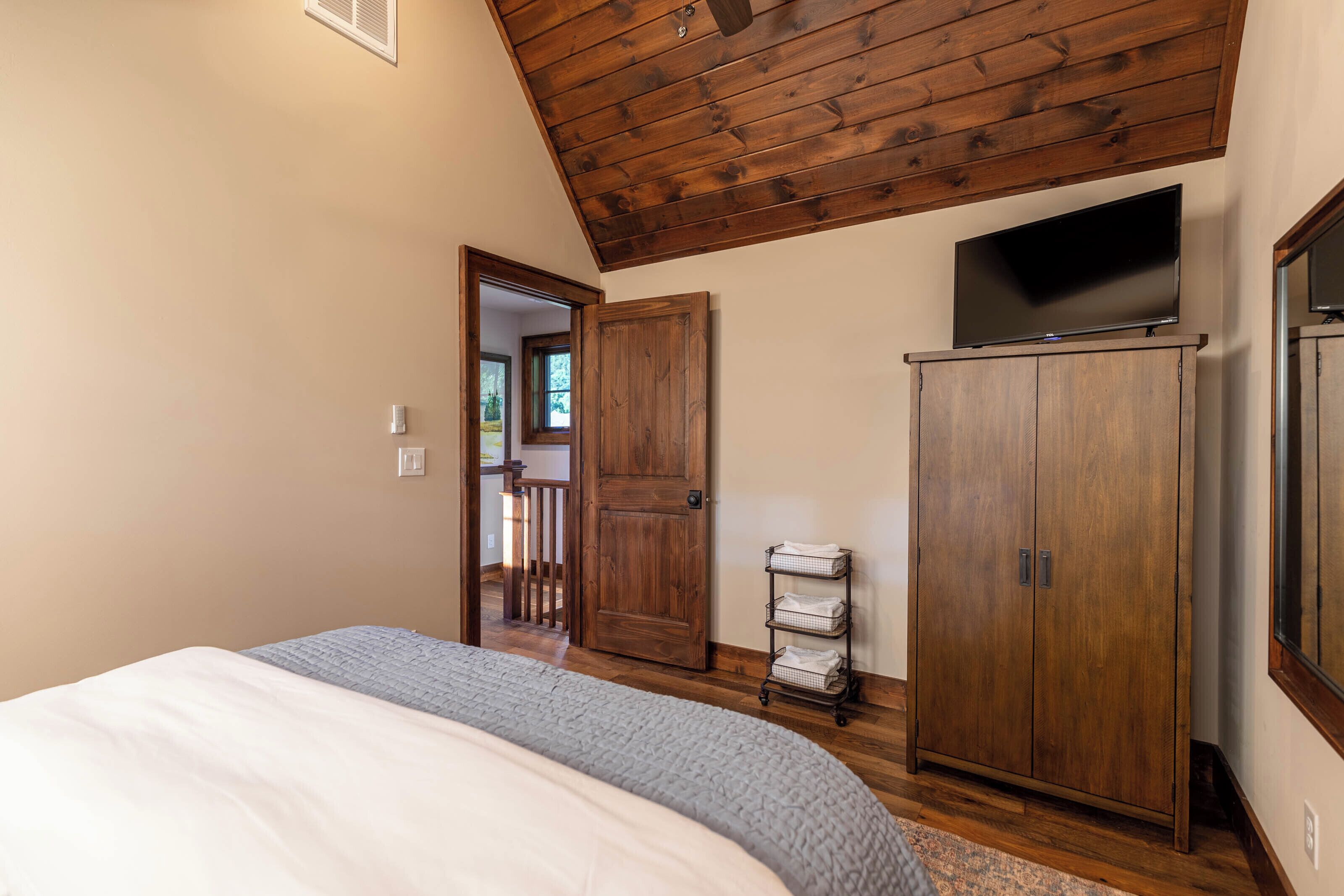Un dormitorio con techo de madera, una cama con manta gris, un armario de madera y un televisor encima.