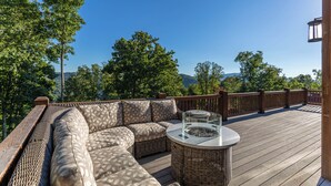Terraza de madera con sofá de mimbre y una mesa con superficie de vidrio, rodeada por barandilla de madera.