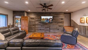 A spacious living room with a large leather sofa, a blue recliner, a wooden coffee table, and a colorful rug. A flat-screen TV is mounted on a stone wall, and there is a staircase leading to another level. - Boulder Sky at River Run (Banner Elk)