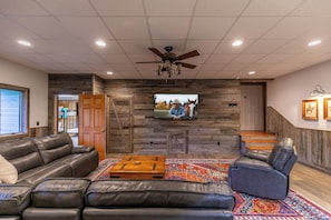 A spacious living room with a large leather sofa, a blue recliner, a wooden coffee table, and a colorful rug. A flat-screen TV is mounted on a stone wall, and there is a staircase leading to another level. - Boulder Sky at River Run (Banner Elk)