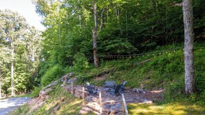 A wooden bench and chairs by a forested area with a stone pathway leading up. - Boulder Sky at River Run (Banner Elk)