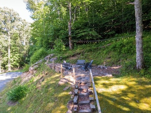 A wooden bench and chairs by a forested area with a stone pathway leading up. - Boulder Sky at River Run (Banner Elk)