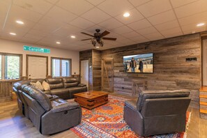 A spacious living room with a sectional sofa, leather armchairs, a wooden coffee table, and a large television mounted on a wooden wall.