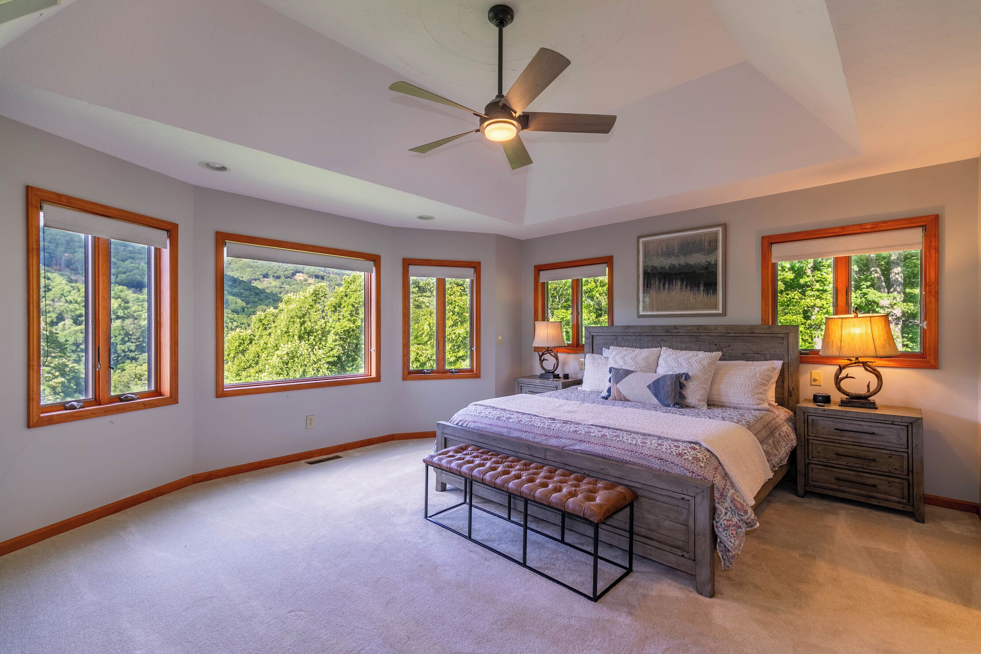 Camera da letto con un letto grande, una panca, una ventola a soffitto e diverse finestre con vista sul verde.