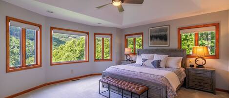 A bedroom with a large bed, a bench, a ceiling fan, and multiple windows offering a view of greenery.