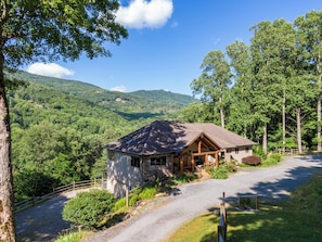 A rustic cabin with a stone foundation, wooden exterior, and a sloped roof, surrounded by lush greenery and hills in the background.