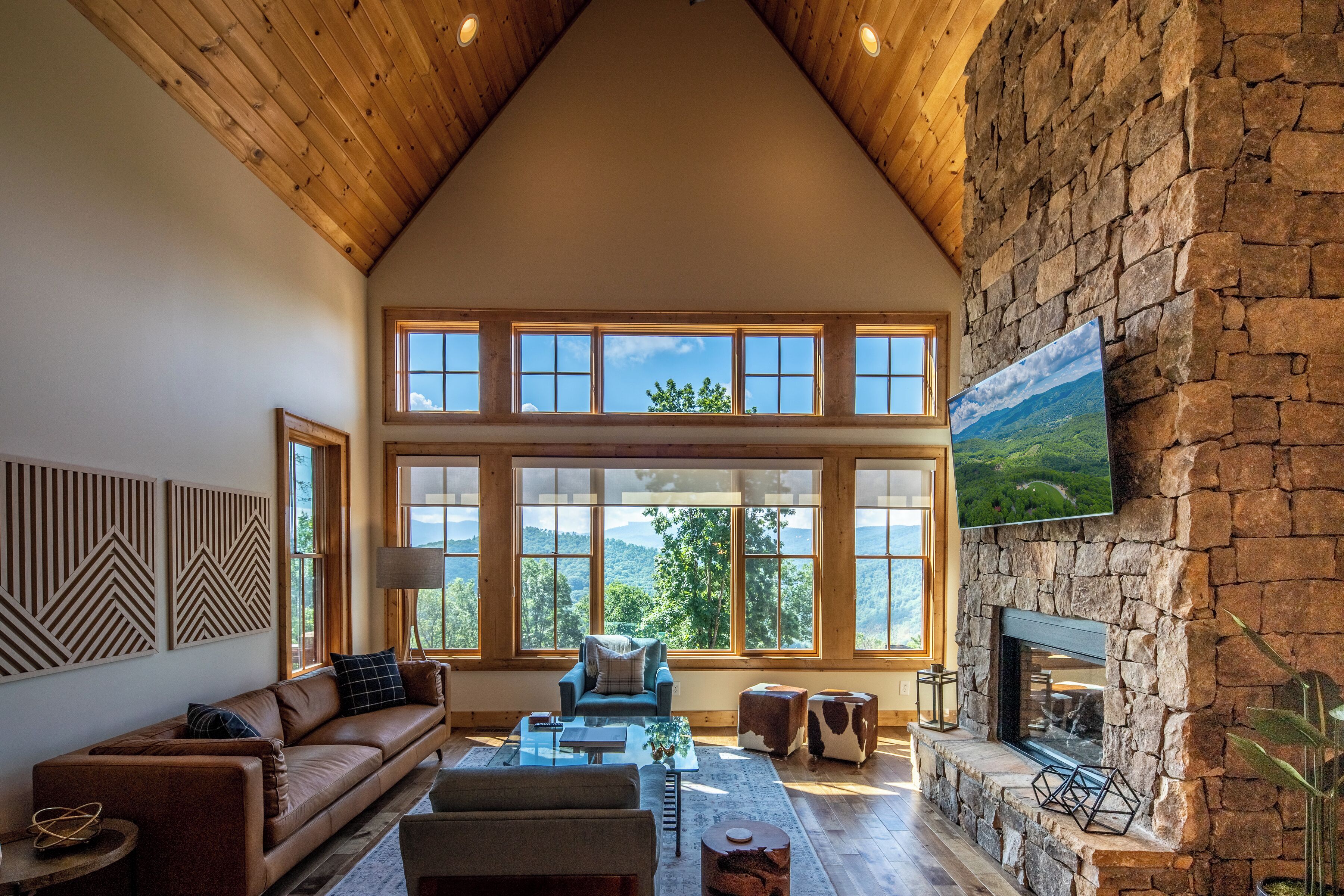 A spacious living room with a high ceiling, large windows, a stone fireplace, and a mounted television.