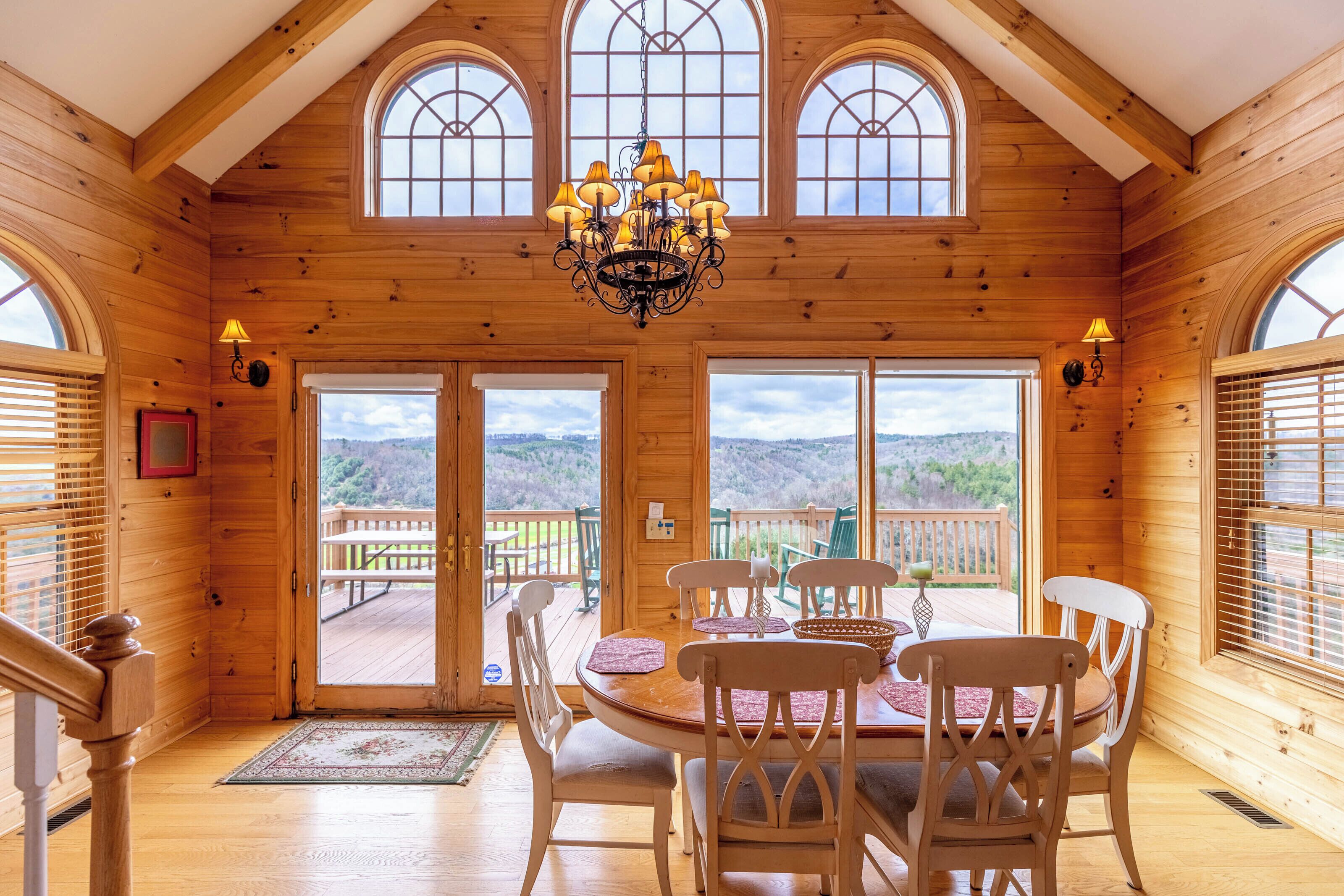 A wooden interior with a dining table, chairs, chandelier, and large windows.