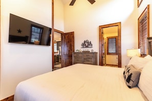 Quarto com cama, cômoda, televisão fixada na parede e porta que dá para outro espaço.